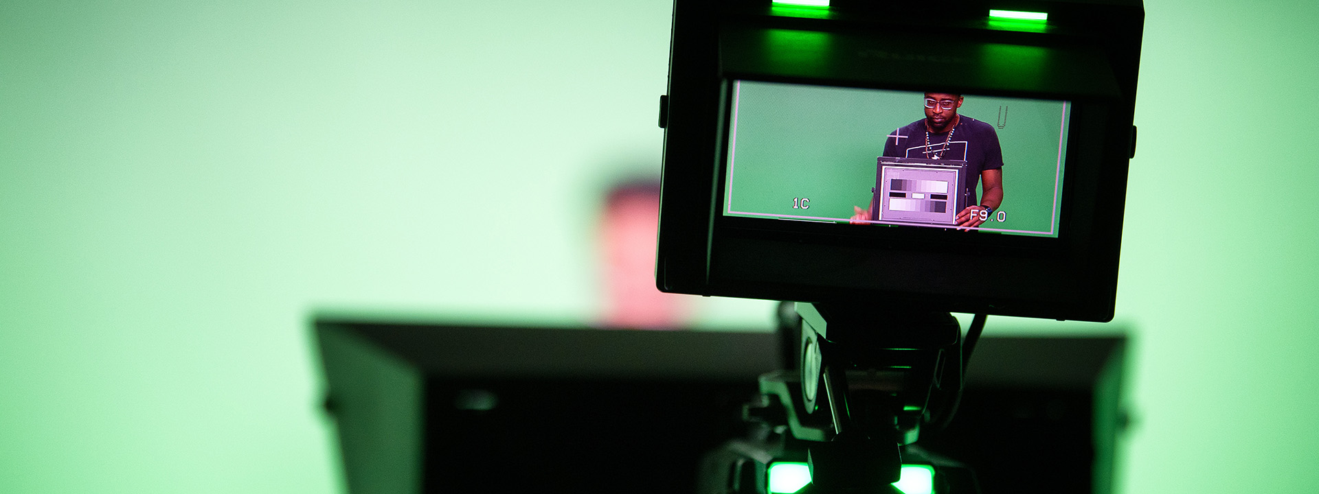 a student in a broadcast studio with green screen is seen through a camera display