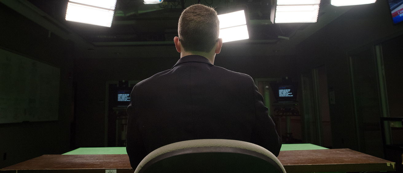 a view from behind a broadcaster sitting at a desk in a studio, where lights from various angles are shining down on him