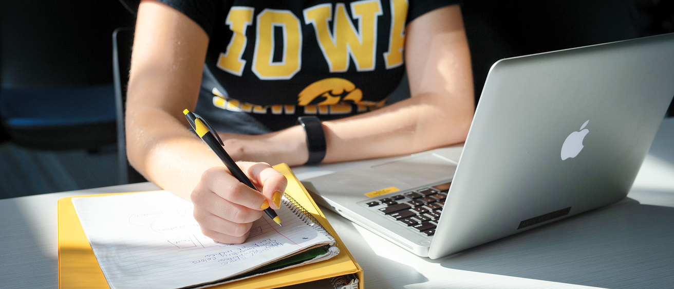 a person studying at a desk, writing in a notebook, with a laptop on the desk nearby