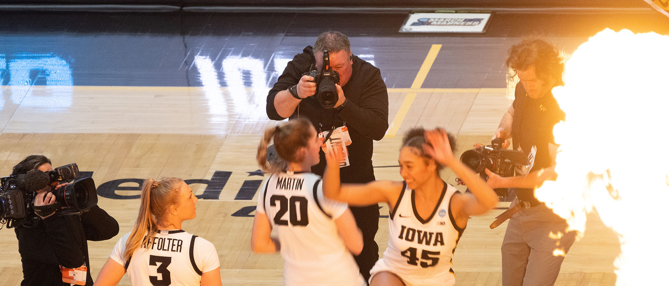 a photographer working at a college basketball game, shooting photos of two players hip-checking each other during lineup announcements