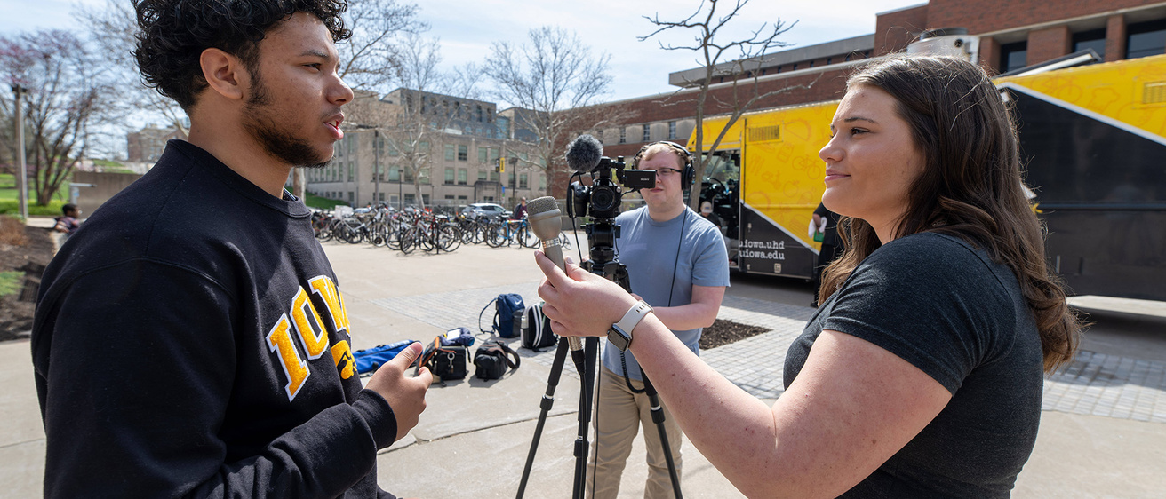 an interview on a college campus, where a woman asks a man questions, while a second man operates a video camera
