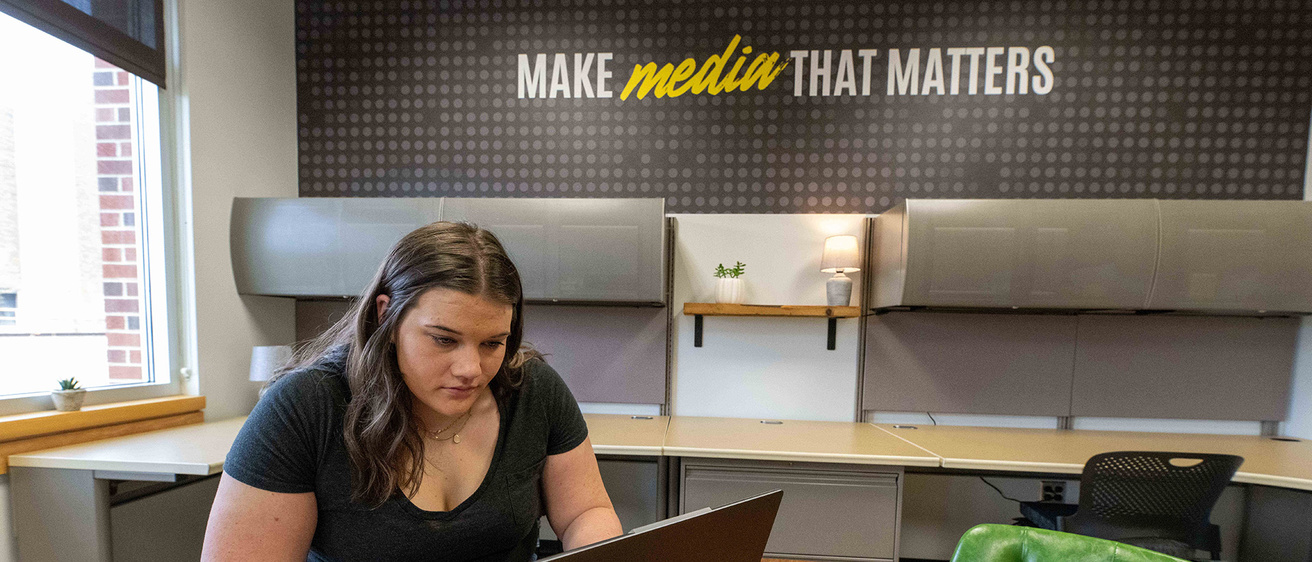 a woman works on a laptop in a room with the phrase "make media that matters" written on the wall in the background