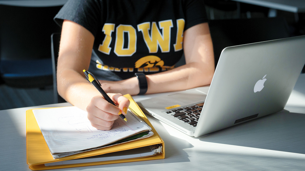 a person studying at a desk, writing in a notebook, with a laptop on the desk nearby