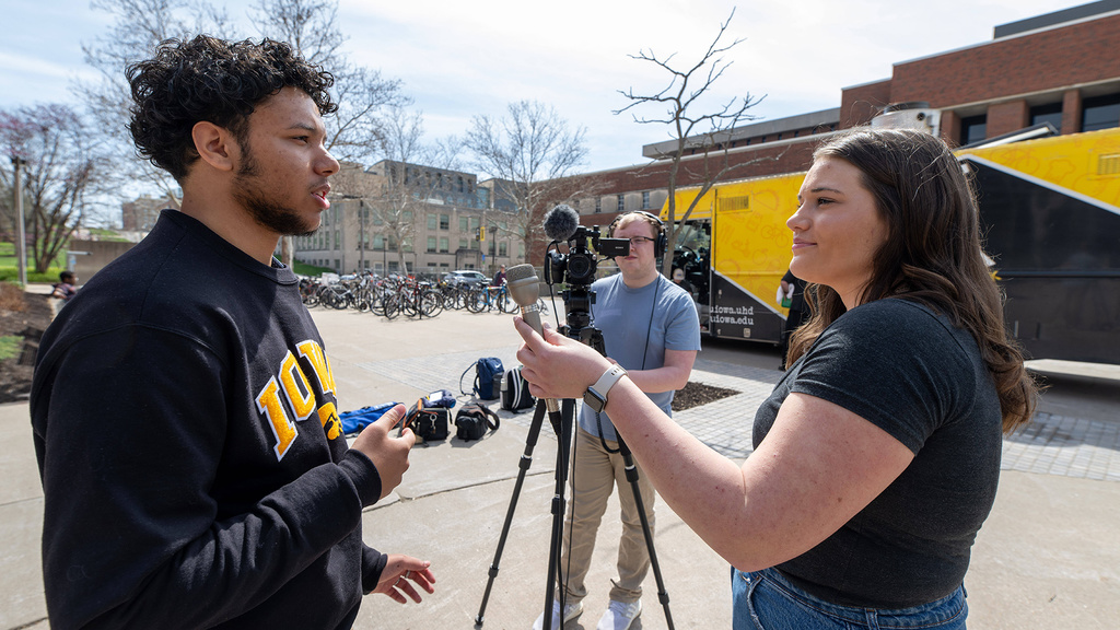 an interview on a college campus, where a woman asks a man questions, while a second man operates a video camera